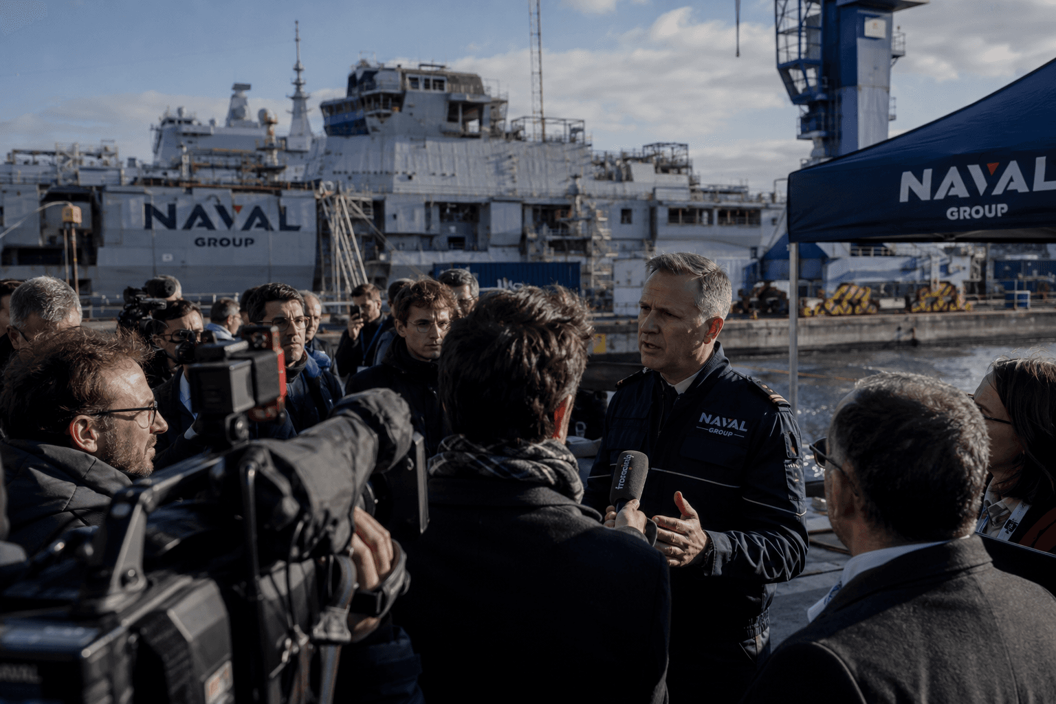 Point de presse sur un quai de chantier naval, un porte-parole en interview face aux journalistes et cameras, coque de navire en arriere-plan