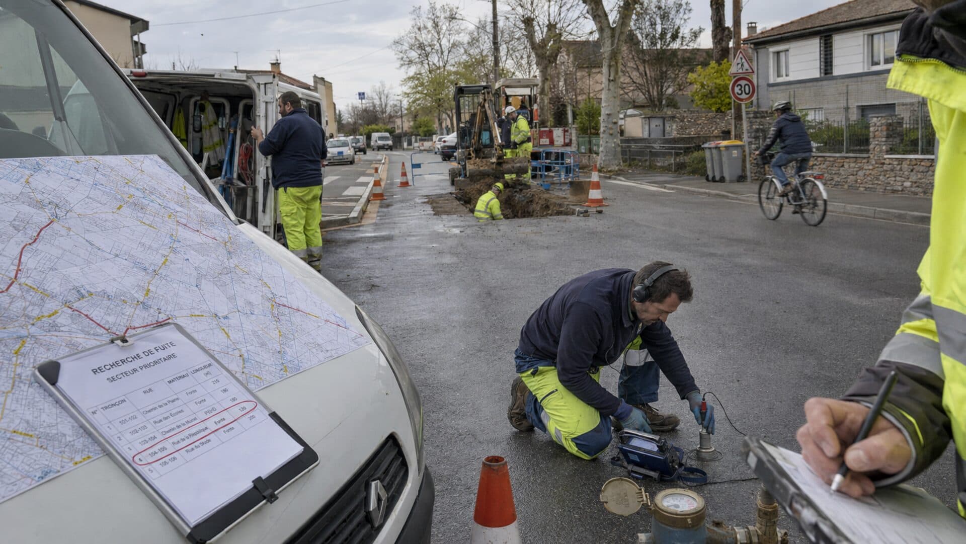 Deux agents municipaux en recherche de fuite dans une rue résidentielle : plan du réseau posé sur le fourgon, technicien à l'écoute acoustique agenouillé, relevé sur le carnet, chantier de réparation en arrière-plan