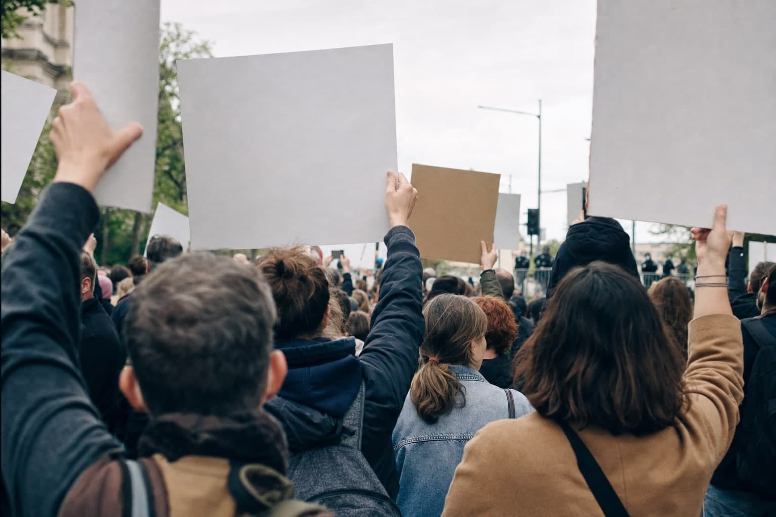 En plein jour sur une avenue, une manifestation pacifique photographiee depuis l'interieur du cortege a hauteur d'epaule, des dos et des bras leves au premier plan, des pancartes vierges brandies au-dessus de la foule et une barriere metallique au fond, aucun visage identifiable