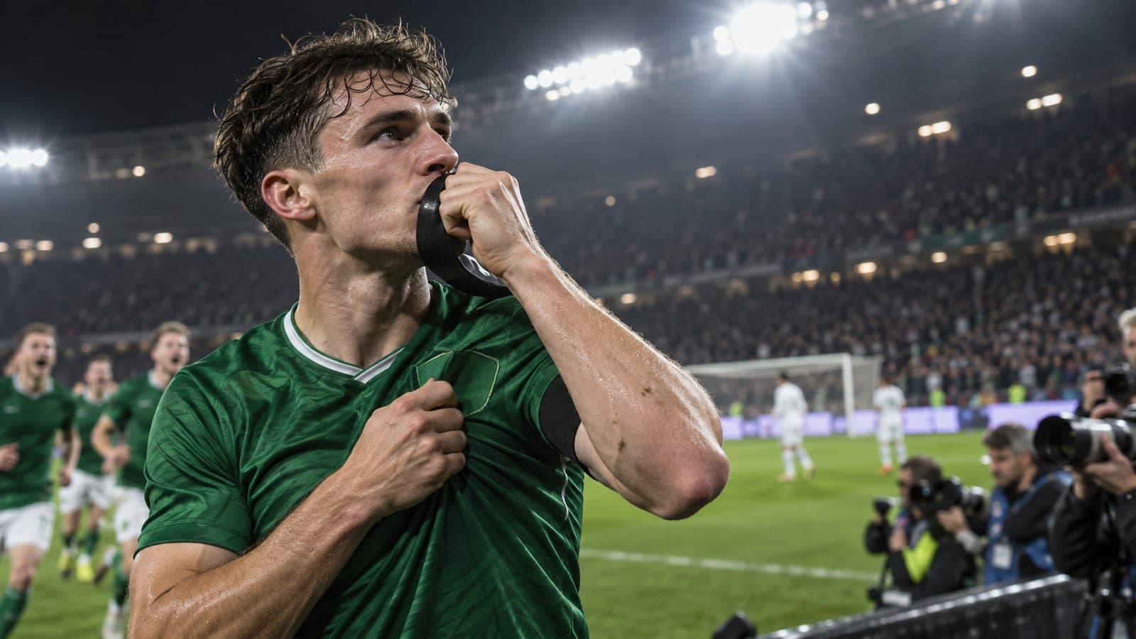 Footballeur en maillot vert portant un brassard noir au bras, embrassant ce brassard et saisissant l'écusson de son maillot en célébrant, dans un stade éclairé de nuit, photographes de presse au premier plan