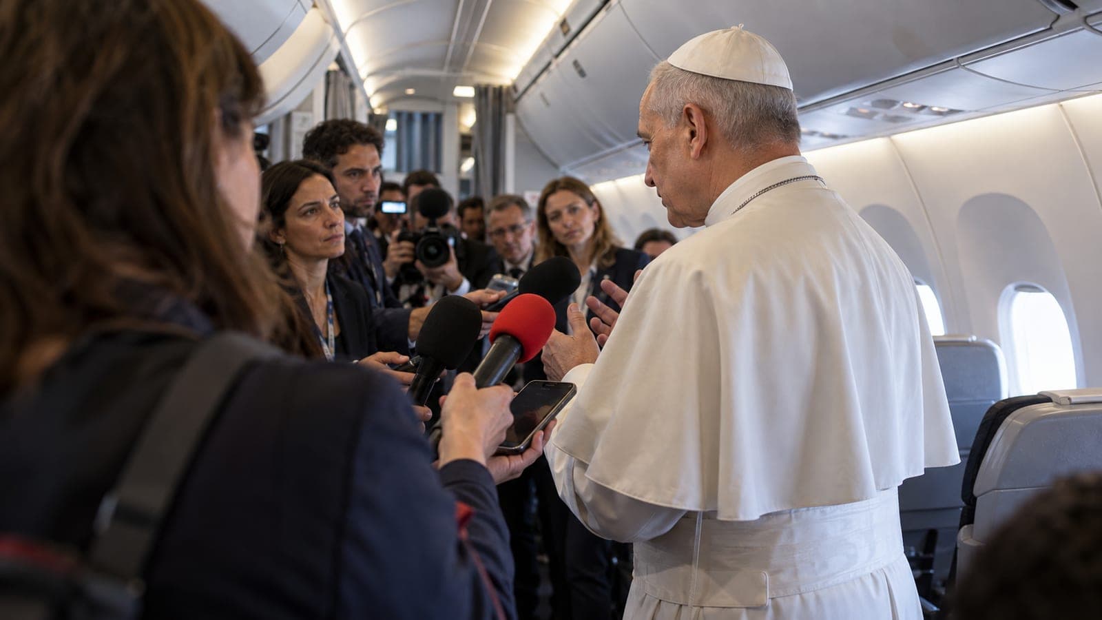 Illustration éditoriale : à bord d'un avion, un homme en soutane blanche répond à un groupe de journalistes qui lui tendent micros et téléphones dans l'allée centrale