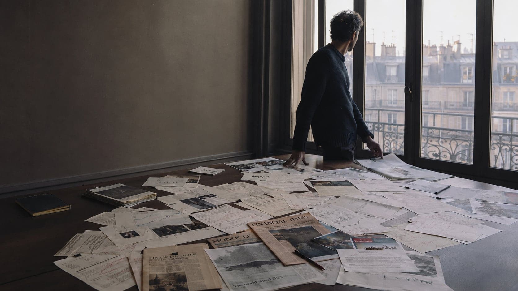 Un stratège observe Paris depuis une fenêtre haussmannienne, une grande table couverte de coupures de presse et de notes annotées derrière lui.