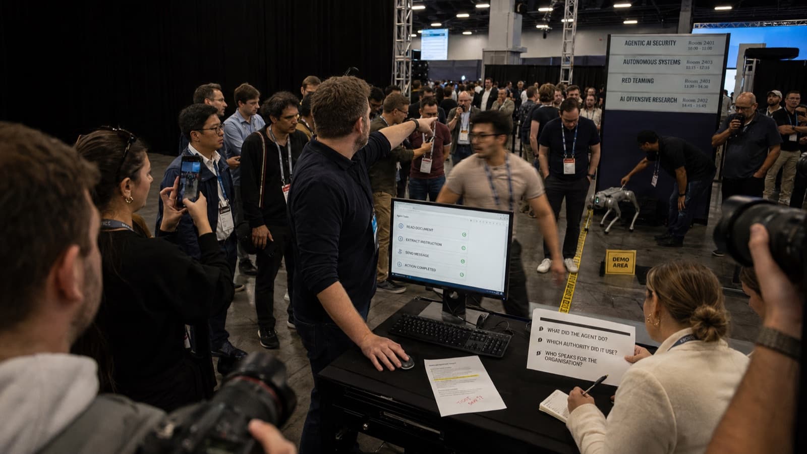 Sur le salon Black Hat, une démonstration publique de détournement d'agent devant un écran affichant les étapes lire le document, extraire l'instruction, envoyer le message, action terminée. Au premier plan, une participante note trois questions, qu'a fait l'agent, quelle autorisation a-t-il utilisée, qui parle au nom de l'organisation. Illustration de la crise déclenchée par une décision automatisée, charte ELMARQ.