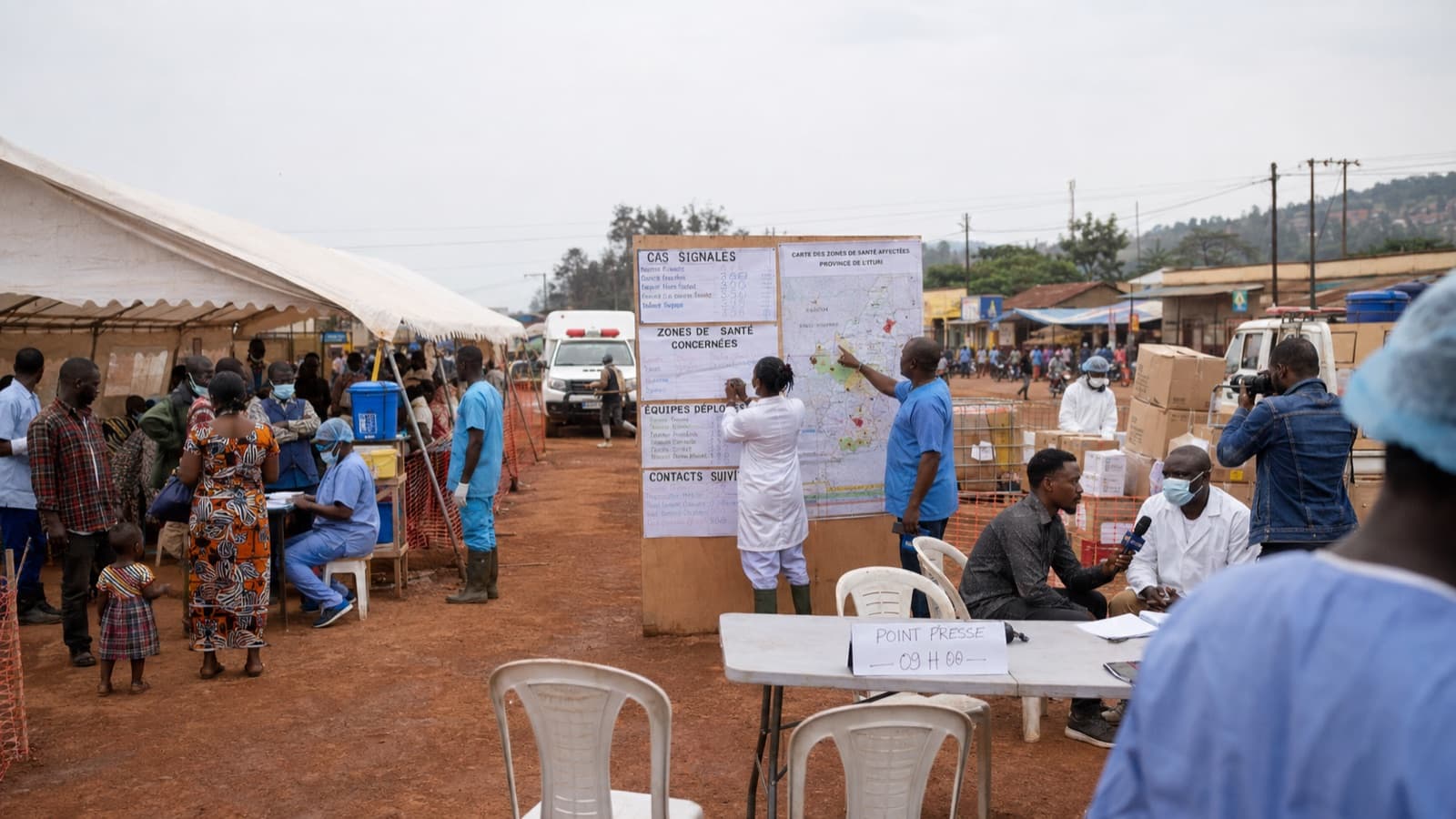Sur un site de riposte en Ituri, des soignants tiennent à jour un tableau des cas signalés, des zones de santé concernées et des équipes déployées, à côté d'une carte des zones affectées, pendant qu'un point presse se tient à une table voisine. Illustration du travail de terrain qui reste hors du champ médiatique, charte ELMARQ.