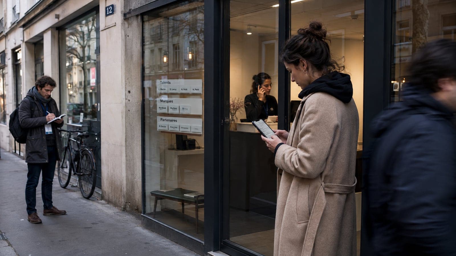 Devant la devanture d'une agence d'influenceurs affichant ses talents, ses marques et ses partenaires, une passante consulte son téléphone tandis qu'un journaliste prend des notes sur le trottoir, illustrant la chaîne d'imputation qui relie une personne, son agence et ses partenaires