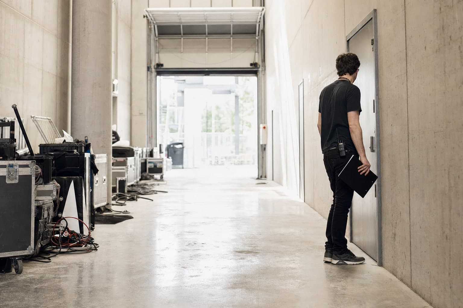 En milieu de journee, dans le couloir technique betonne qui longe l'arriere d'une scene de stade, des caisses de transport alignees et des cables au sol, un regisseur planchette a la main se tient immobile devant une porte metallique fermee, la lumiere du jour entrant par un quai releve au fond