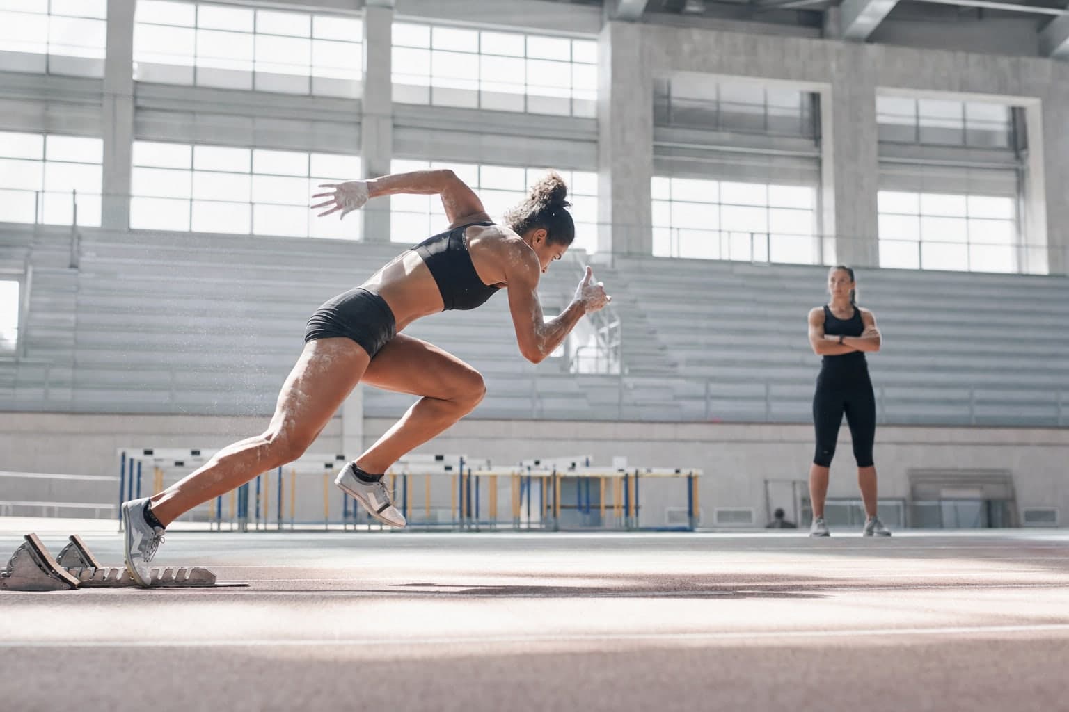 En milieu de matinee sur une piste d'athletisme couverte inondee de lumiere, une athlete en plein effort sort des blocs de depart, muscles tendus et sueur visible, tandis qu'une entraineuse chronometre au bord du couloir, gradins vides derriere