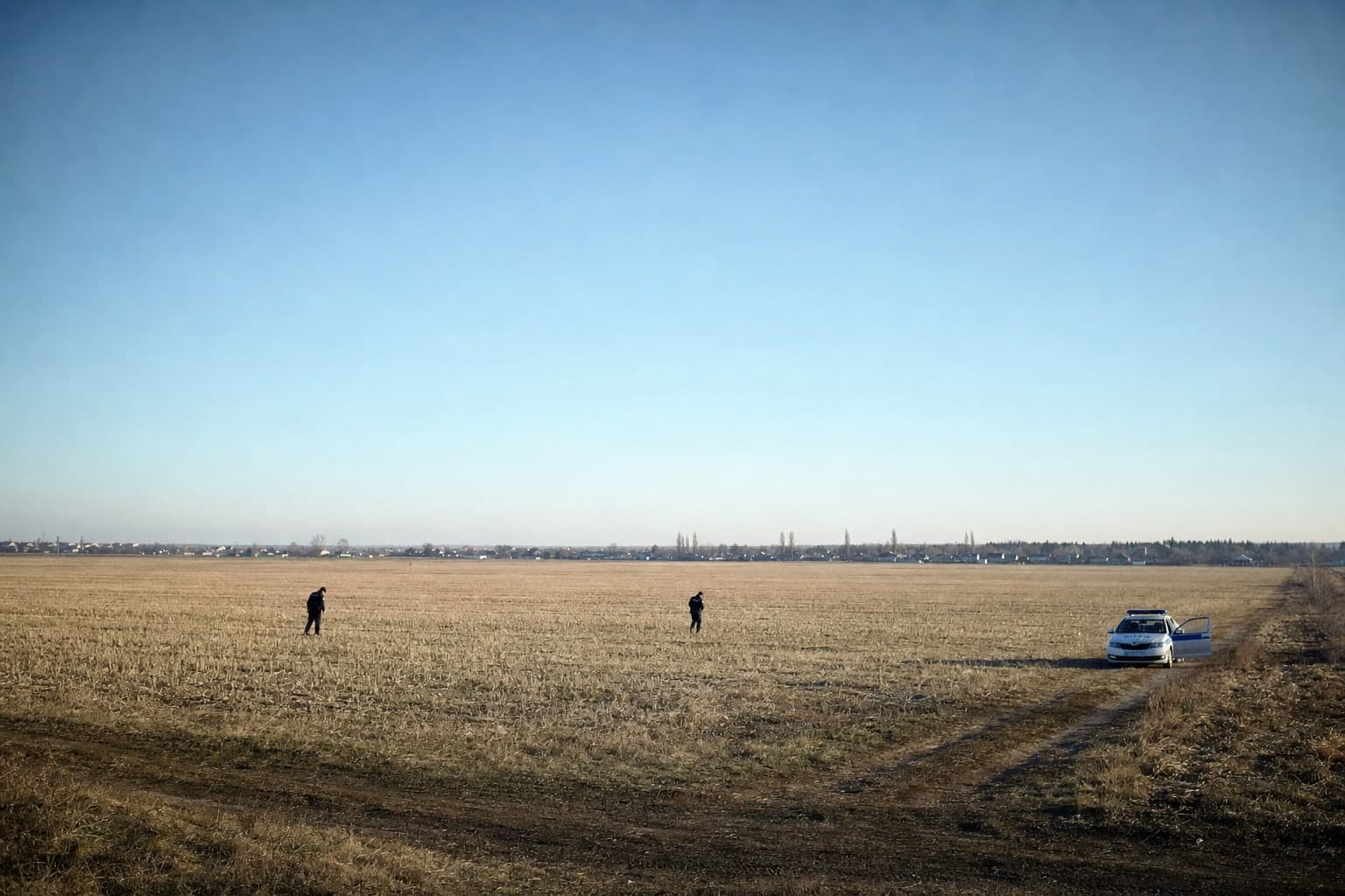 Petit matin dans un champ de chaume sous un ciel entierement vide, 2 policiers marchent separement tete baissee en scrutant le sol, une voiture de patrouille garee au bord du champ portiere ouverte et une ligne de village a l'horizon, illustrant une recherche qui ne trouve rien