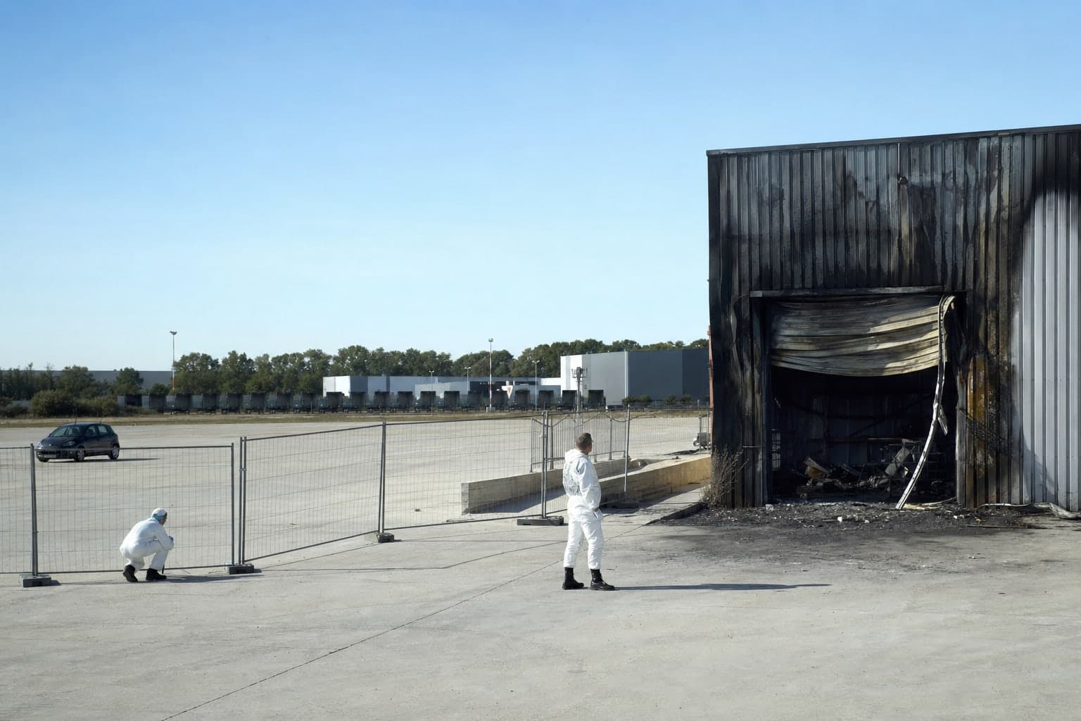 En plein jour sous un ciel clair, 2 enqueteurs en combinaison marchent le long d'une cloture provisoire devant un box de stockage industriel a la tole noircie par un incendie, sur une vaste aire de beton pale en bordure d'une zone logistique, l'un accroupi examinant le sol et l'autre observant la structure