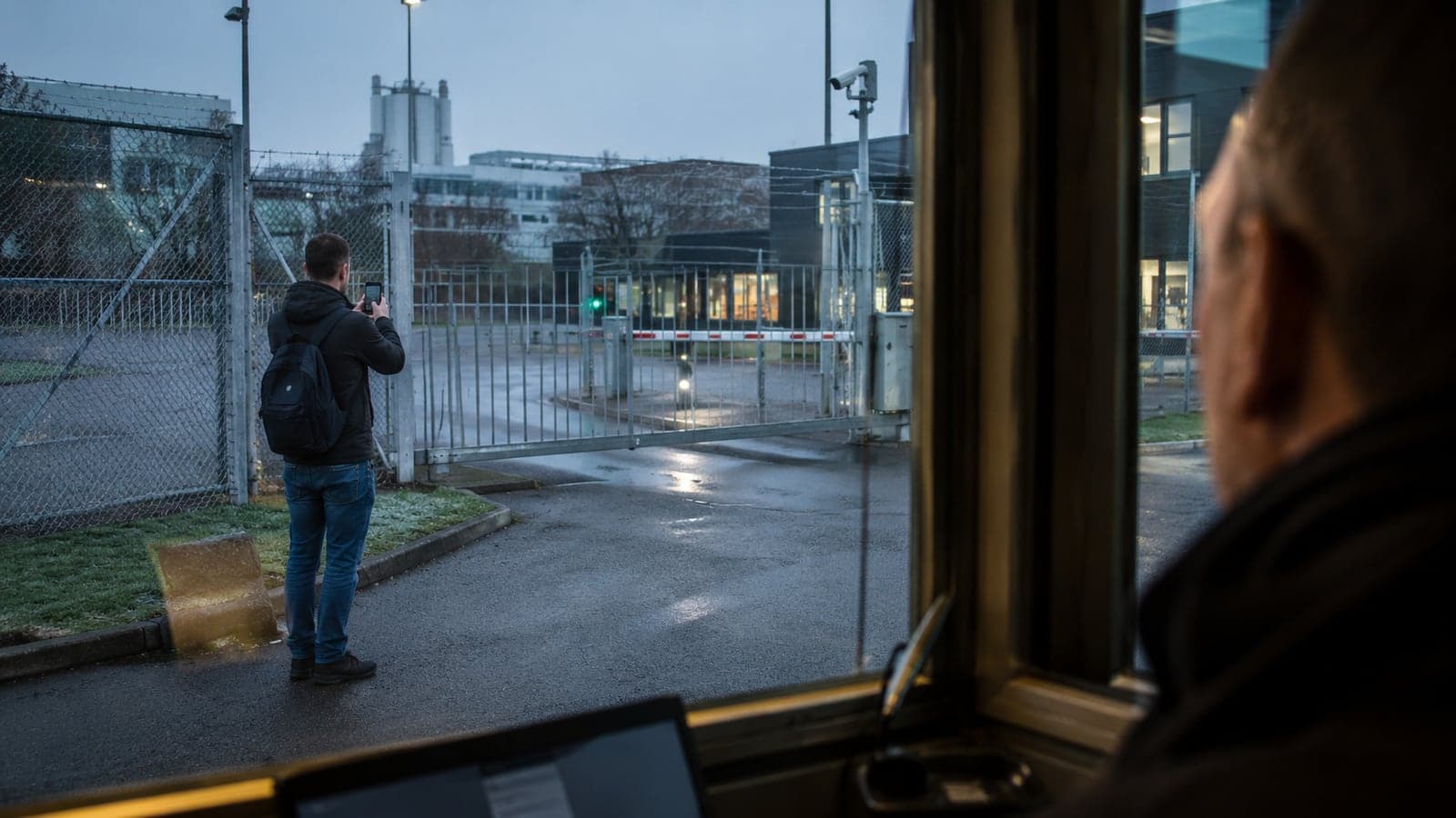À l'aube, devant la grille et la barrière d'un site industriel, un homme au sac à dos photographie l'entrée avec son téléphone, observé depuis la guérite par un agent de sécurité, illustration d'une reconnaissance anodine confiée à un citoyen ordinaire.
