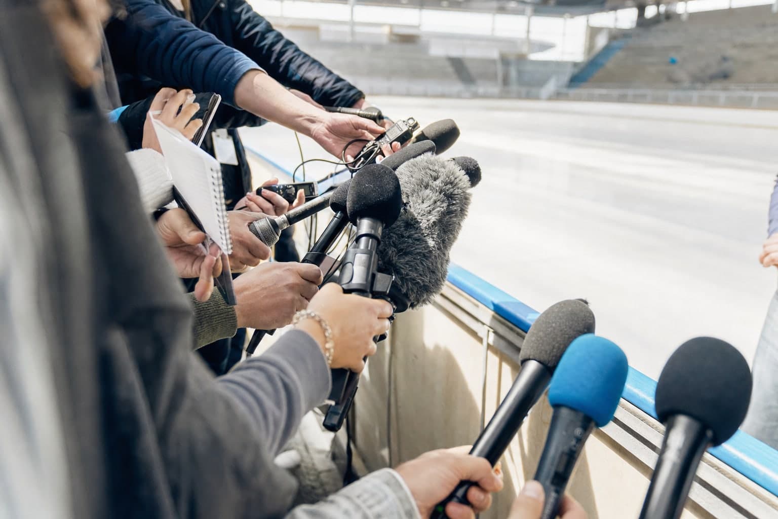 Lors d'une journee presse dans une patinoire d'entrainement baignee de lumiere, une grappe dense de micros et d'enregistreurs est tendue vers un interlocuteur hors champ par une dizaine de journalistes debout en demi-cercle, gradins vides et glace claire en arriere-plan
